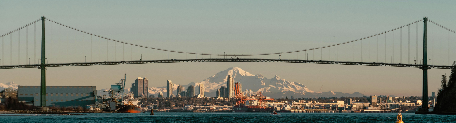 Scenic view of Lions Gate suspension bridge that connects cities, and in the background Burrard inlet full of cranes and industrial harbor, with Mount Baker appearing behind. 