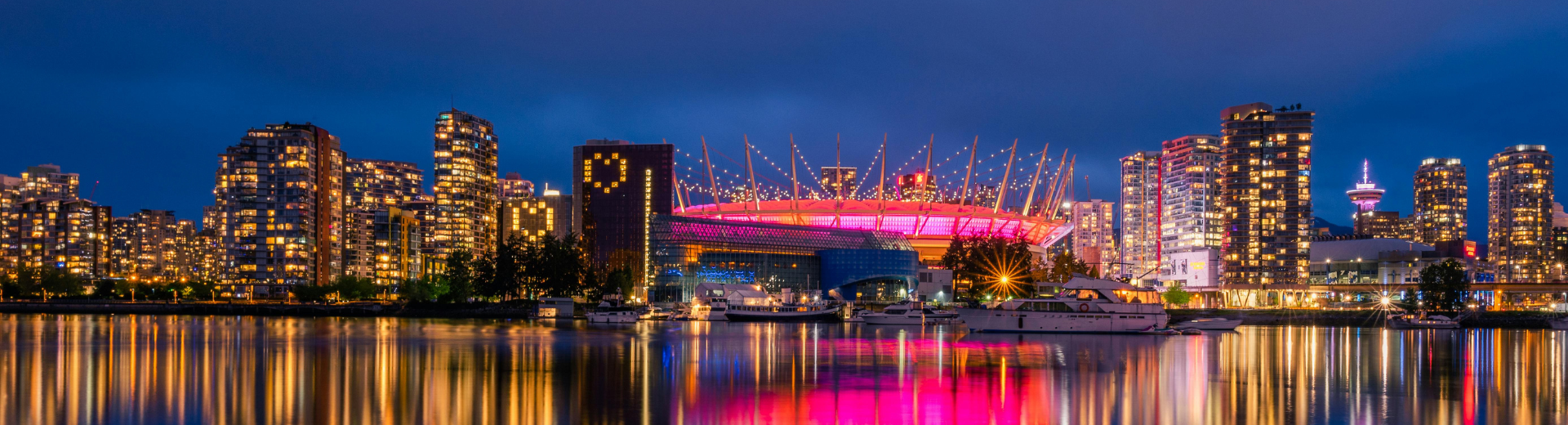 A night view of Downtown Vancouver skyline, where BC place is highlighted due to its magenta lights, all high-rise buildings are lightened up, and the lights of the city reflects on the sea water of the waterfront